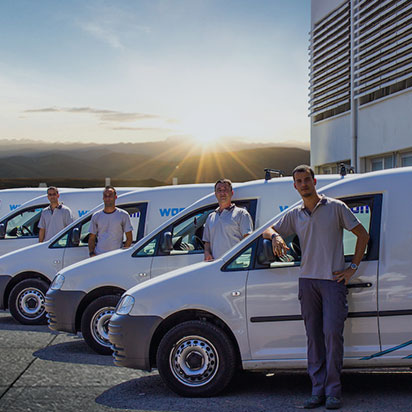 Quatro profissionais de uniforme ao lado de quatro carrinhas de serviço Wondercom brancas ao nascer do sol, com um edifício à direita e montanhas ao fundo.