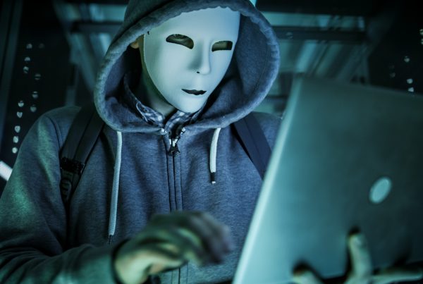 Close-up Shot of a Masked Hacker in a Hoodie Standing in the Middle of Data Center full of Rack Servers and Hacking it with His Laptop.
