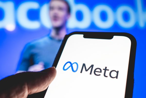 Logotipo da Meta exibido na tela de um smartphone, com Mark Zuckerberg desfocado ao fundo, dando uma apresentação. Atrás dele, pode-se ver "Facebook" escrito em branco num fundo azul.