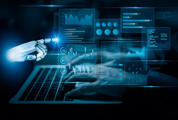 Representação digital de uma mão robótica tocando num ecrã holográfico com diversos gráficos e dados, enquanto mãos humanas digitam num teclado de portátil ao fundo. Ambiente futurista e tecnológico com iluminação azul.