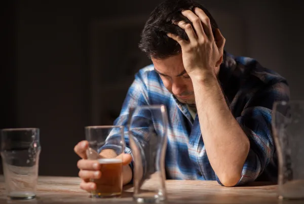 Close-up de um homem sentado numa bancada de madeira com três copos de cerveja à sua frente. Dois copos estão vazios e um está pela metade. O homem aparenta estar triste ou desapontado, com uma mão a cobrir o rosto. A imagem retrata as consequências das adições.
