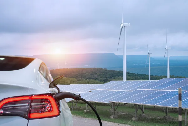 Um carro elétrico branco a ser carregado, com painéis solares e turbinas eólicas ao fundo, numa paisagem natural com montanhas. A imagem destaca a utilização de energias renováveis e a mobilidade sustentável, simbolizando o compromisso com um futuro mais ecológico.